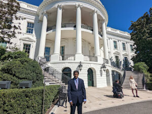 Image shows an outside shot of the White House. Blue sky can be seen in the upper right hand corner and the image it taken looking up at the pillars in the middle of the structure. Hari stands in the grounds below the building, on a concrete path. A man in a wheelchair wearing a black suit, and a women in a cream dress can be seen conversing in the background. 