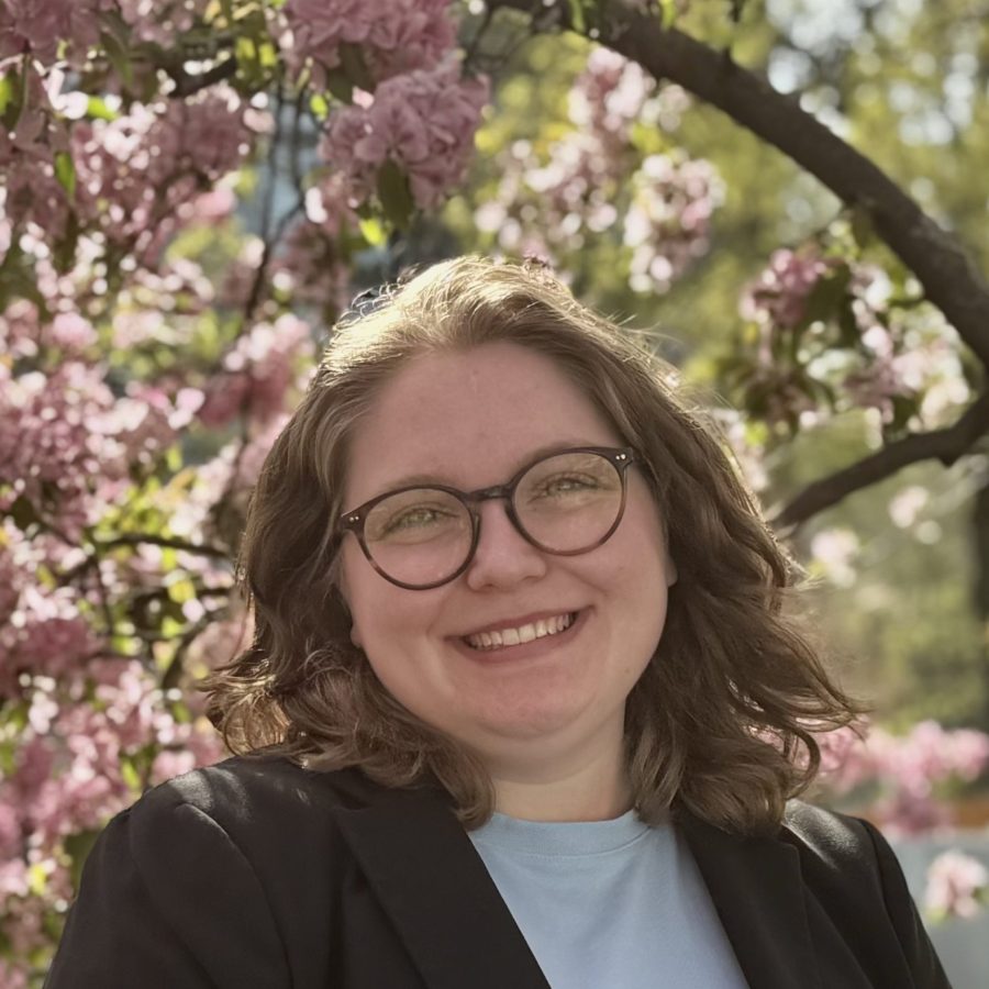 Headshot of white woman with round glasses and shoulder-length brown hair, in light shirt and dark blazer, against leafy background