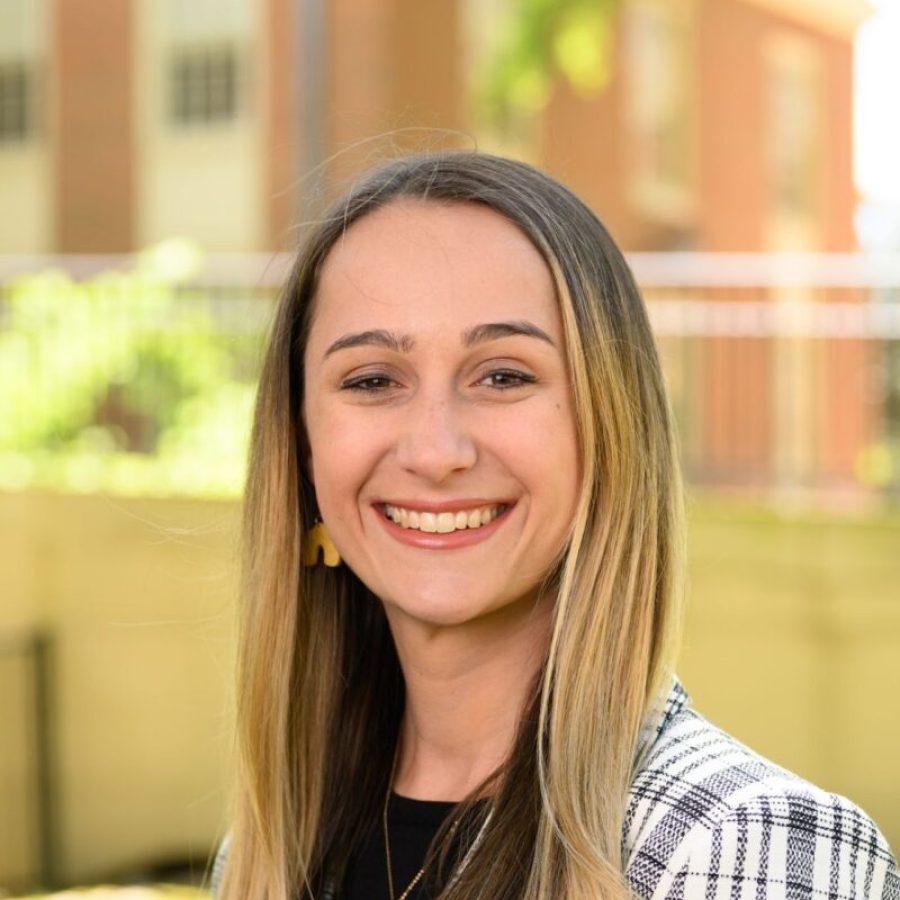 Smiling woman with long blonde hair in black and white blazer and black shirt in front of a brick and stone university building and foliage