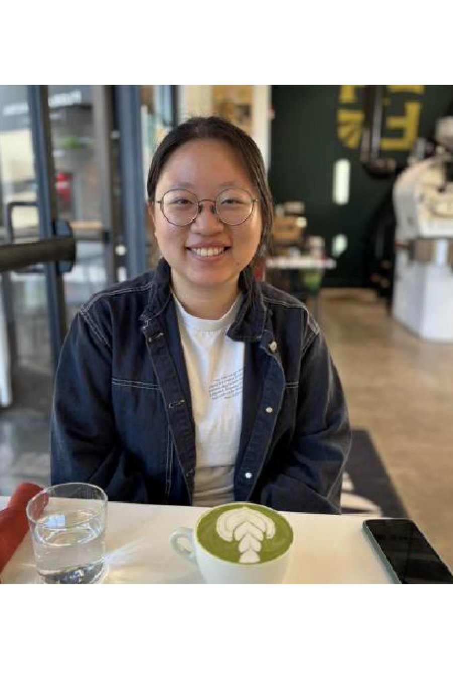 Chinese woman in denim jacket sitting in front of green latte with foam design