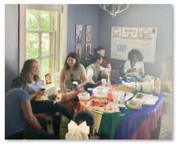 Small groups of students are sitting around a table covered in brunch foods and a rainbow flag.