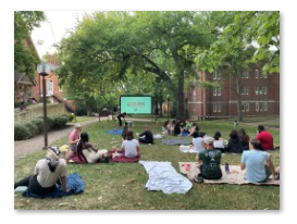 Groups of students are sitting on blankets on the grass. In the background is a large tree and a large projected movie screen with the menu for the movie "But I'm a Cheerleader" shown.