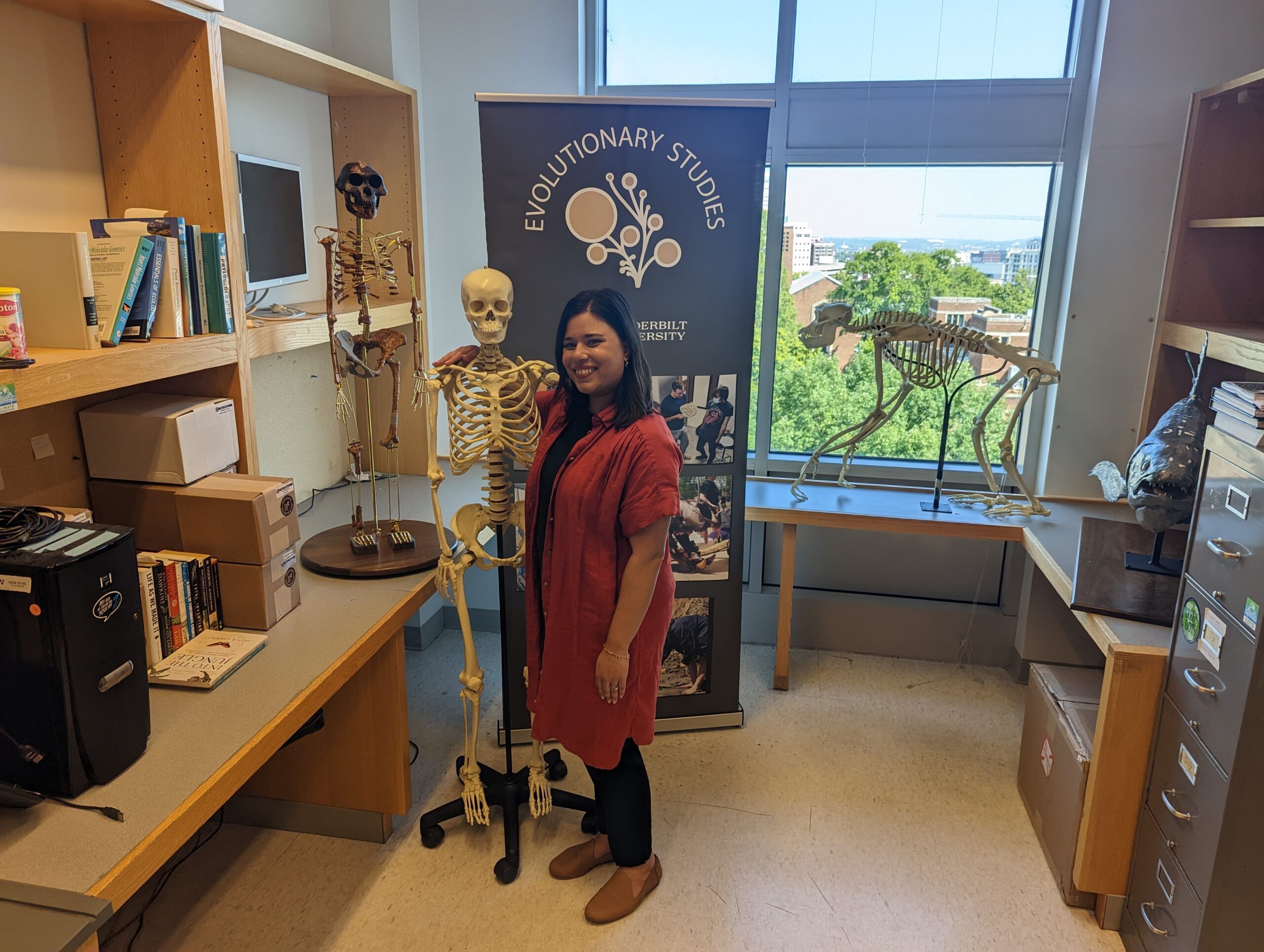 Monica in red standing next to a skeleton cast of a human with a Lucy hominid, chimp, and coelacanth in the background