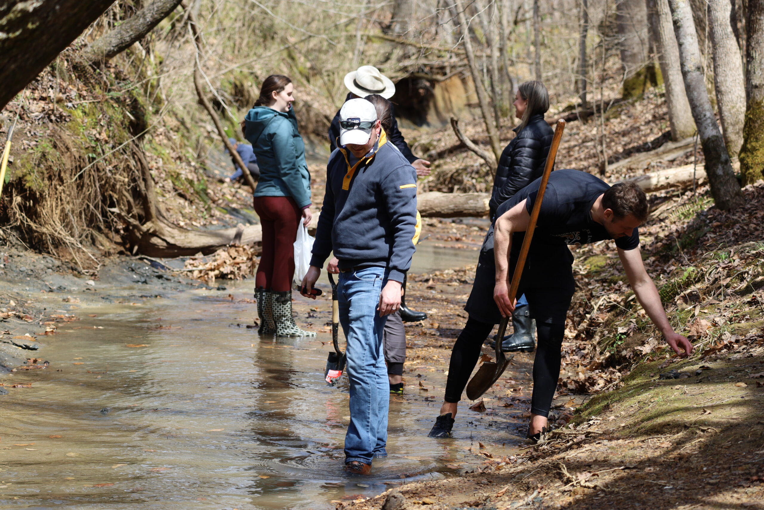 A handful of folks looking at and otherwise inspecting the creek for fossils. Darroch holding a shovel, but not using it, while he leans over to pick up a potential fossil