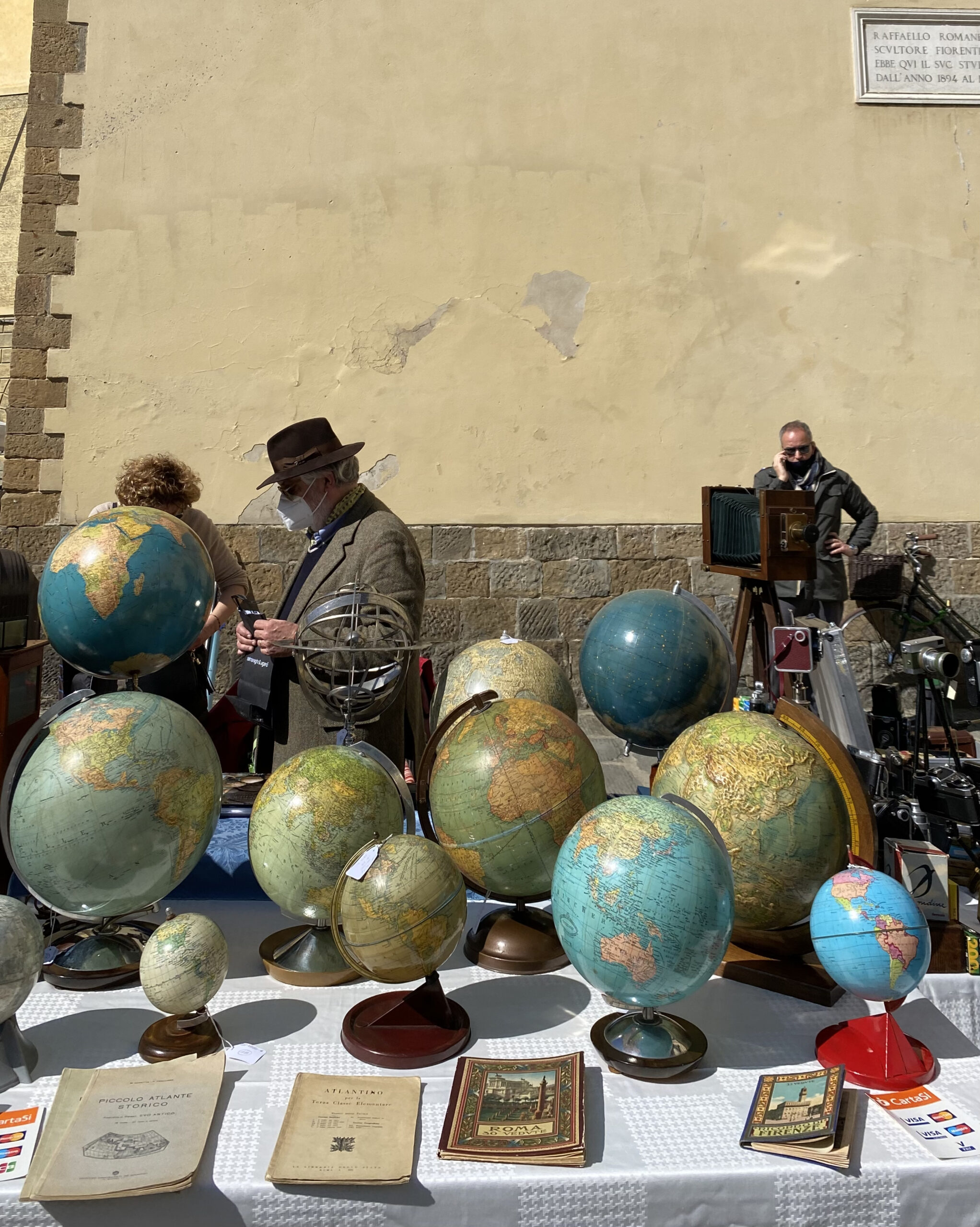 Open-air market stall with globes and antique books, with an artist and shoppers wearing COVID masks in background