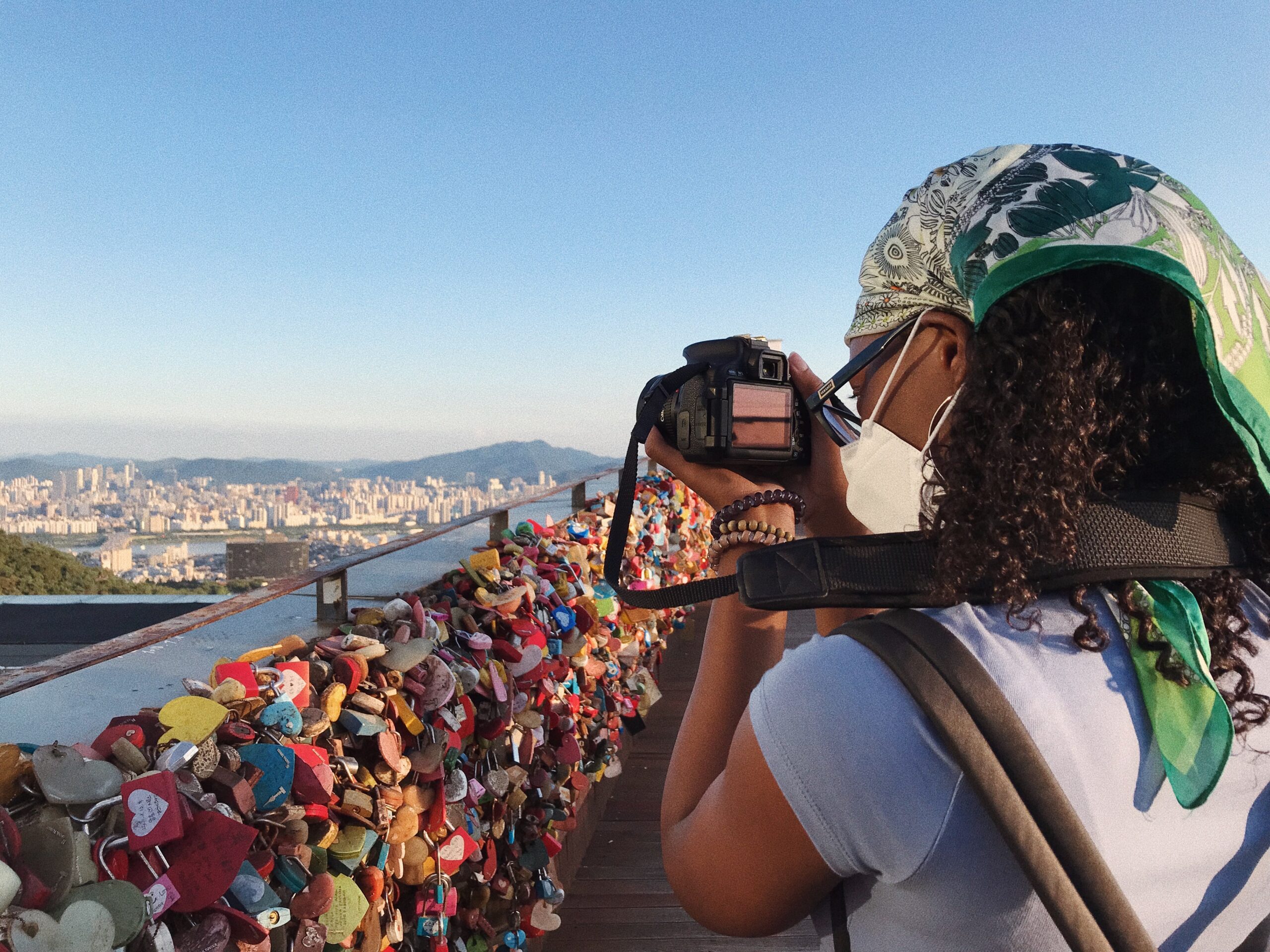 Woman with camera pointed at distant cityscape, with bridge covered in colorful love-locks in foreground