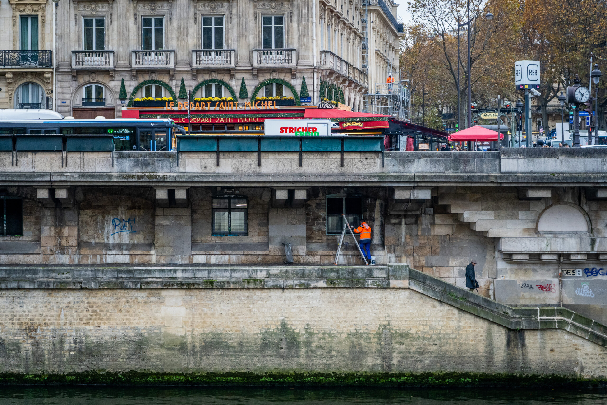Man in an orange jacket working on ladder below street level, above the Seine river
